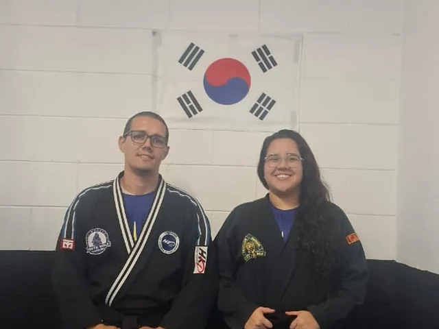 Prof. Henrique Morato com sua assistente, de dobok, sob a bandeira da Coreia no dojô